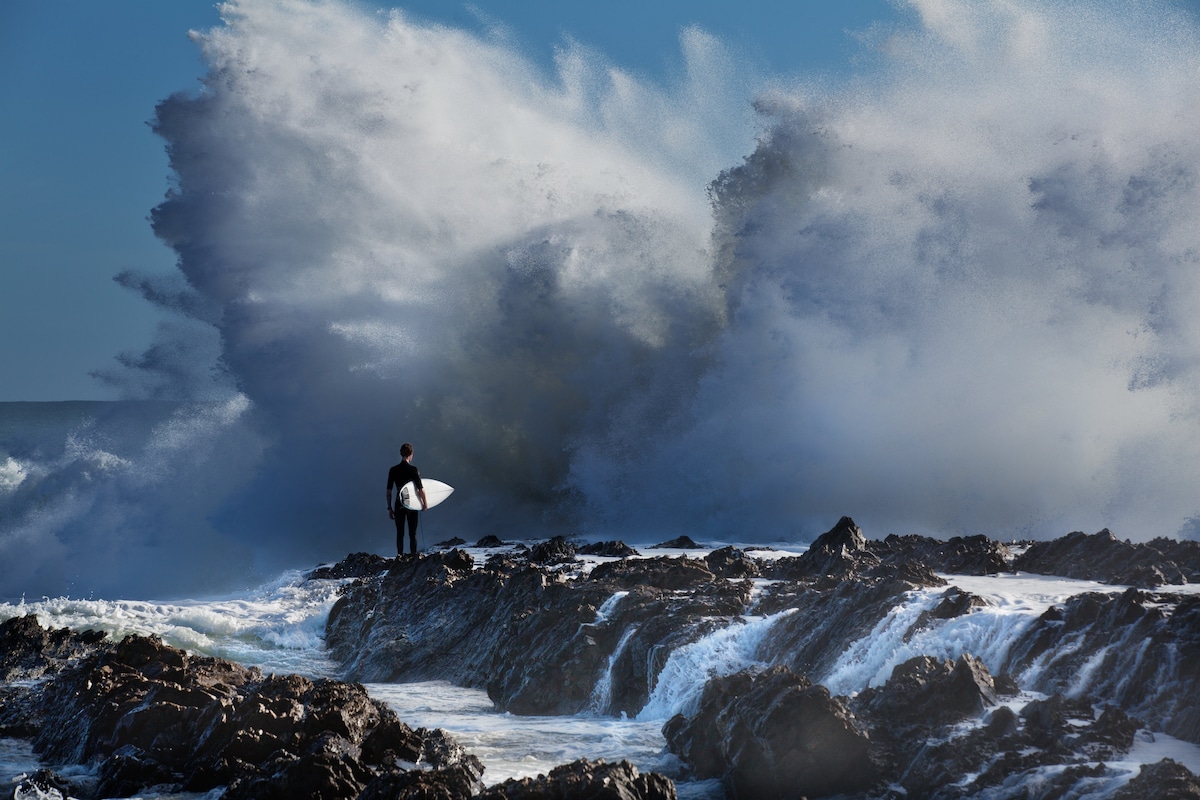 Surfer Standing on Rocks with Waves Crashing