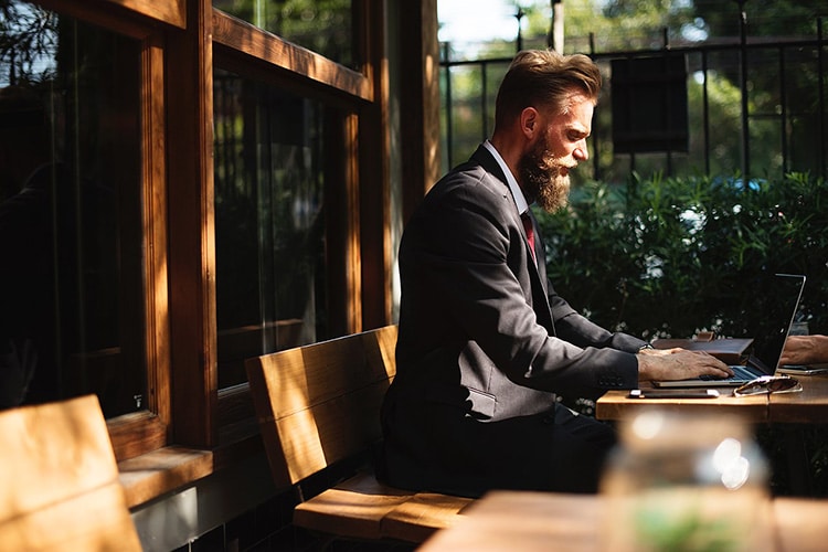 a bearded man in a suit works outside on his laptop surrounded by green flora