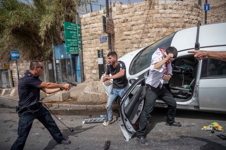 An Israeli police officer stands with his pistol pointed at a Palestinian man, in the center. 