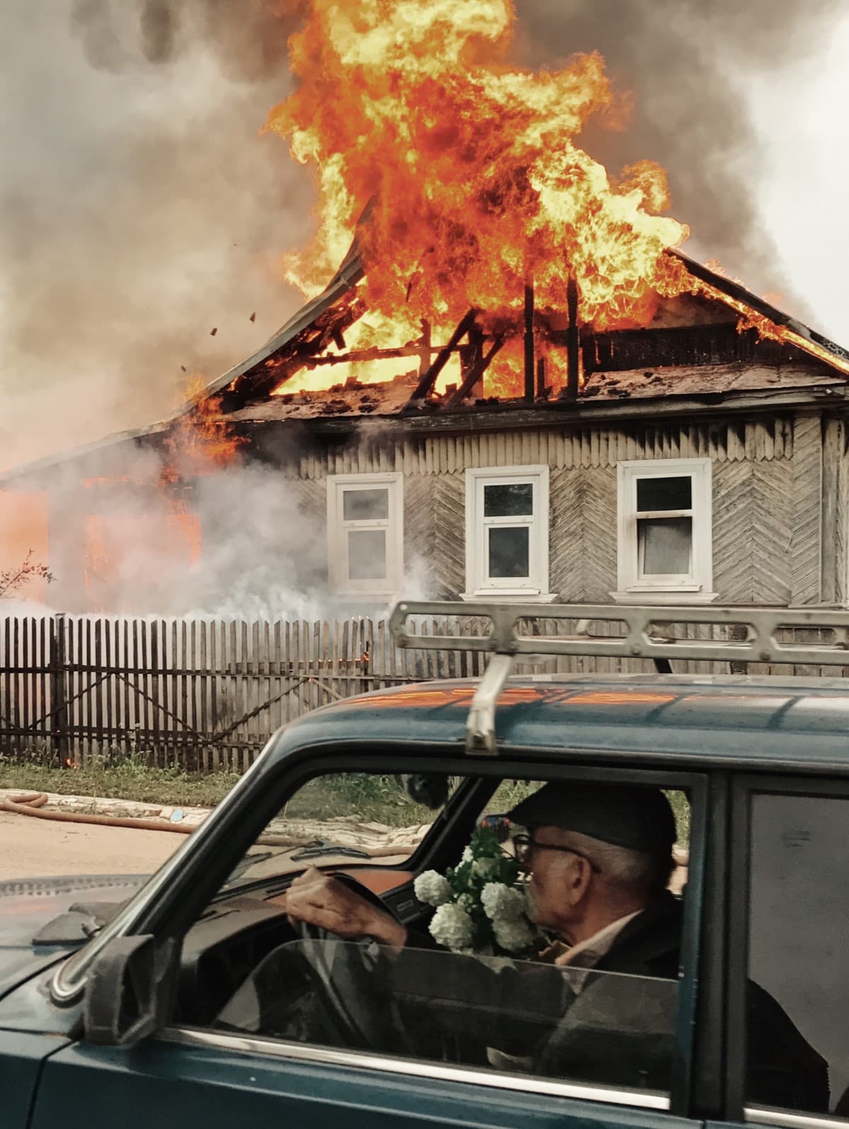 Man sitting in a car outside of a house that's on fire