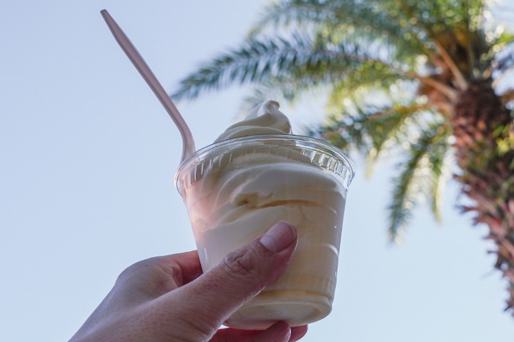 Hand holds up a cup of soft serve pineapple vanilla swirl ice cream, with palm trees in the background