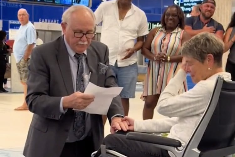 An older man is on his knees as he proposes to girlfriend in an airport