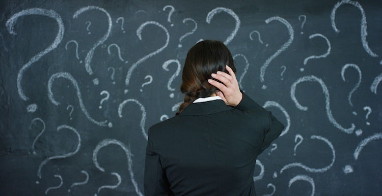 Confused woman with a braid scratching her head, seen from behind. She's standing in front of a chalkboard full of question marks.