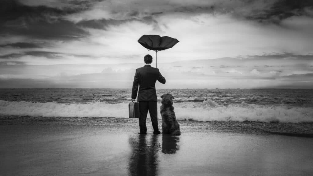 Black and white photo of a man on a beach holding an umbrella with his dog