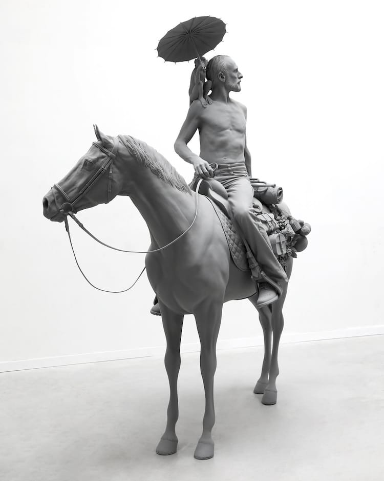 Figurative sculpture of a man on horseback with a monkey holding a parasol on his shoulder in gray monochrome by Hans Op De Beeck