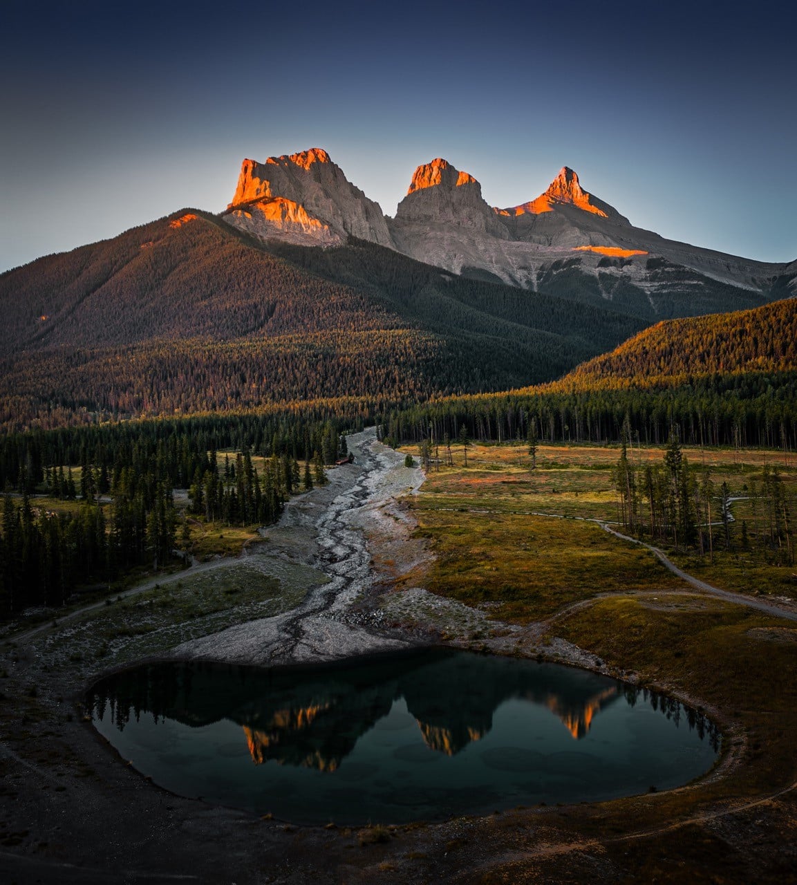 The Three Sisters mountains are reflected in a small lake at sunset in Canmore, Alberta