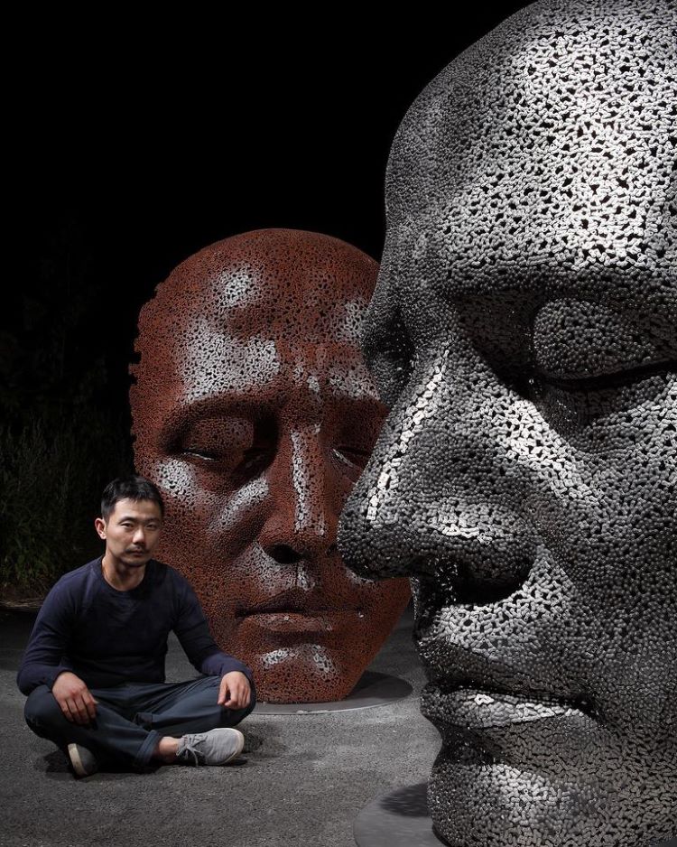 Young Deok Seo Sitting Next To Two Face Sculptures Made Of Bicycle Chains