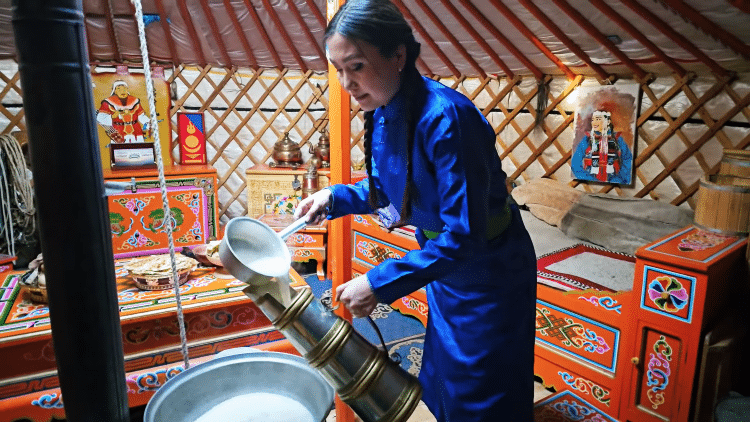 Mongolian Woman In Traditional Blue Dress Spooning Milky Liquid Into Large Drink Container