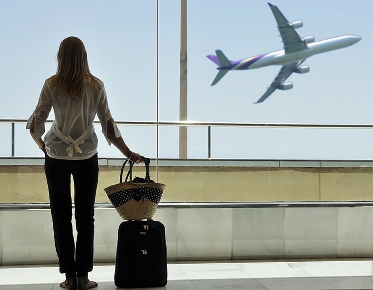 A woman looks out from an airport corridor with her luggage next to her. An airplane takes off in the background.