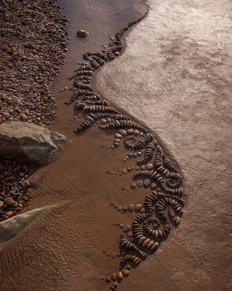 Aqua Exemplaria, 2024 by Jon Foreman is a composition of rocks that mimics the shoreline. This image, shot from a bird's-eye view, features rocks in swirling patterns that extend from the water out towards the sand. 