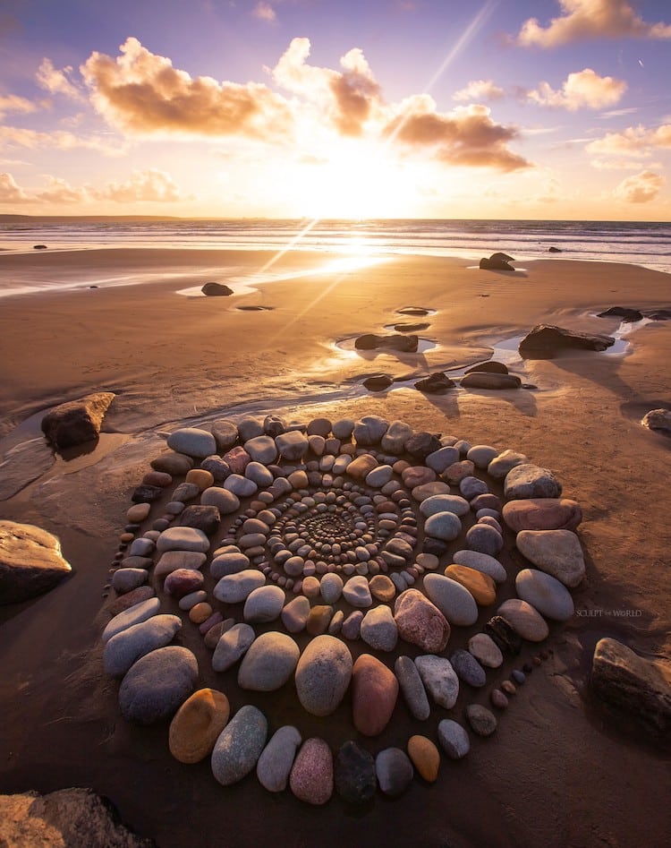 Jon Foreman's Manifold Spiral of 2024 consists of a spiral shape of various stones or rocks on a beach. In the background is the shoreline with the bright sun shining through the clouds. 