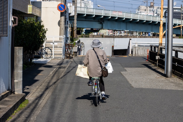Tokyo, Japan. An elderly person cycling on a city street with a bridge and buildings in the background.