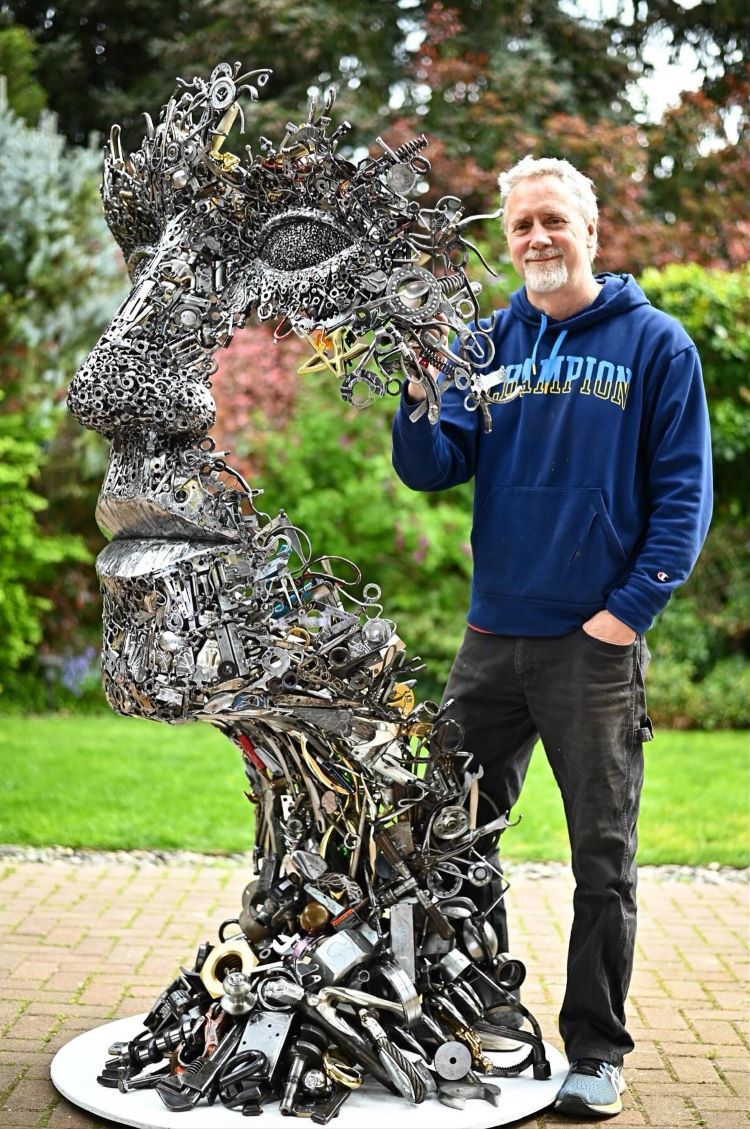 Brian Mock Standing Next To Human Face Sculpture Made Out Of Scrap Metal
