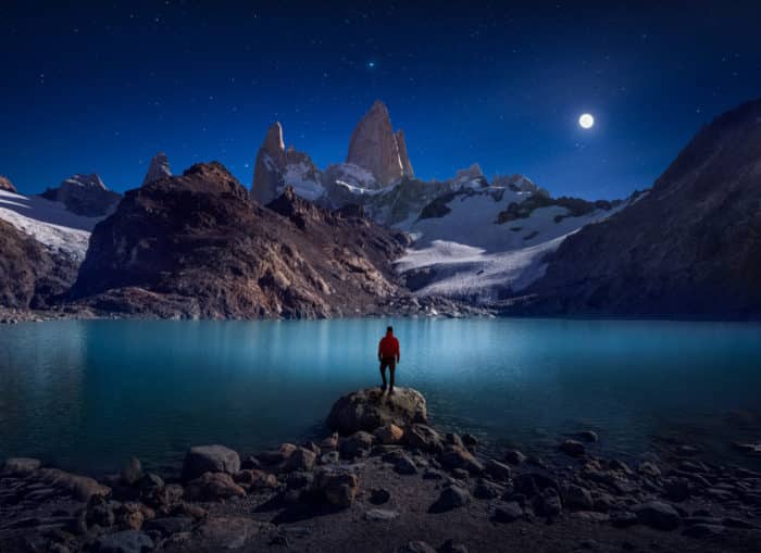 Man standing by a lake at night surrounded by mountains