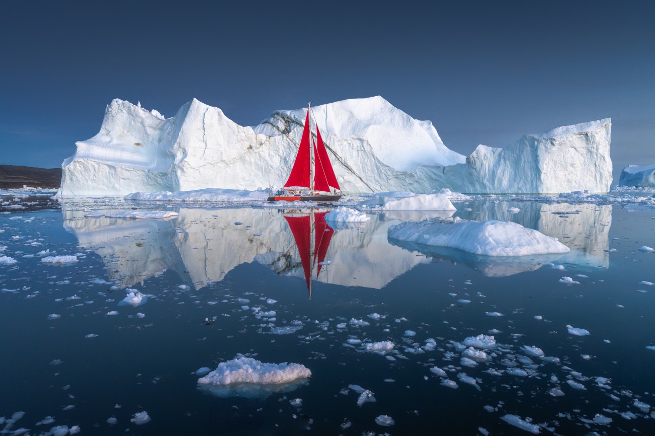 Albert Dros red sailboat in Disco Bay with an iceberg behind it