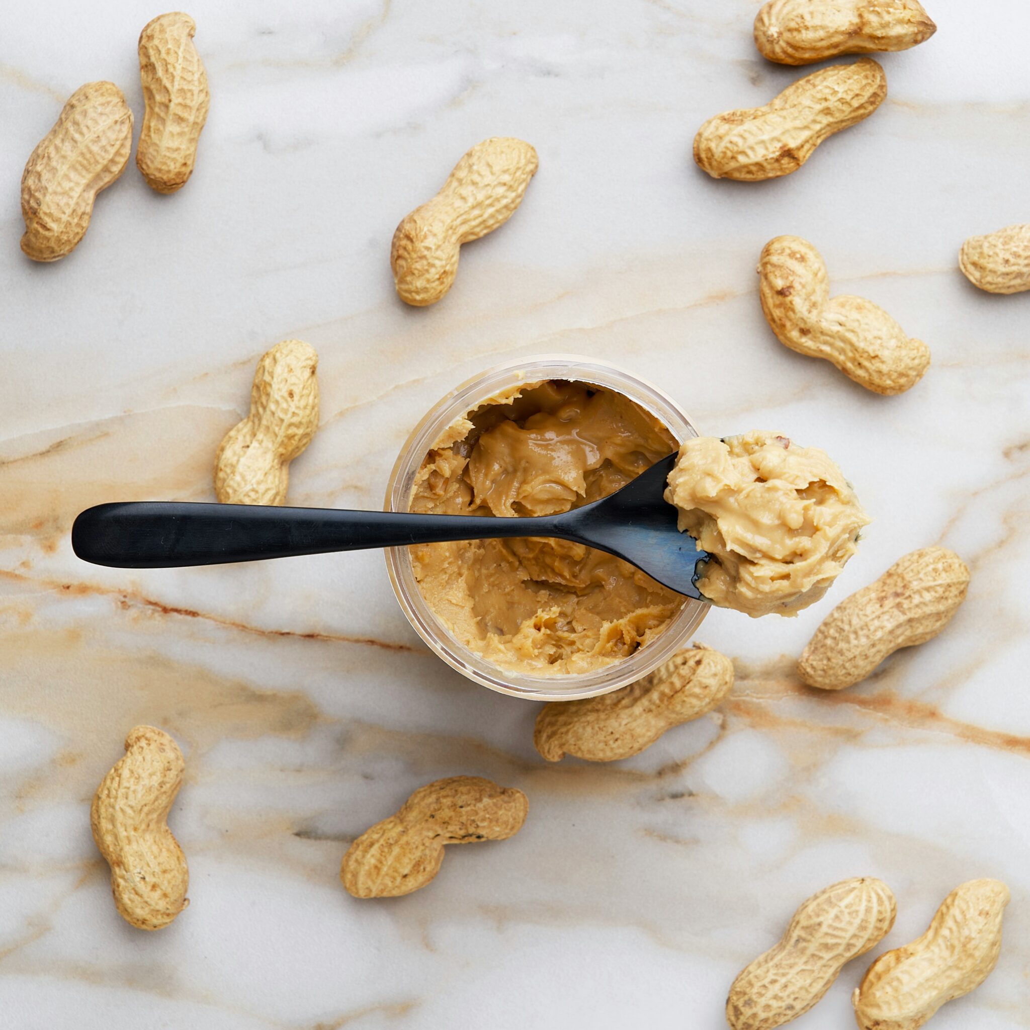 A jar of peanut butter with a spoon resting on top of it. Peanut shells are surrounding the jar, which is placed on a marble table
