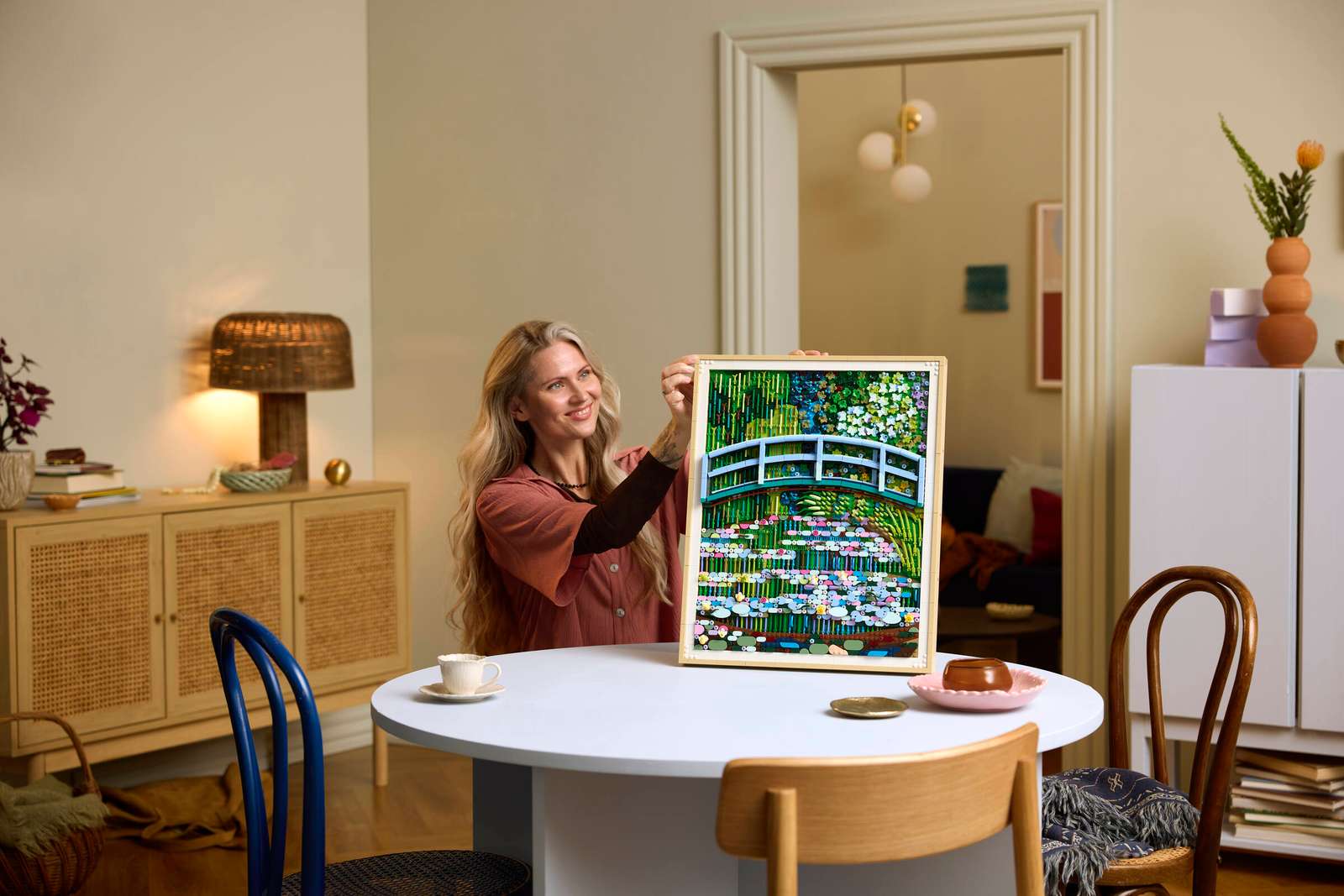 Woman sitting at a table holding up the &lsquo;Monet Bridge Over a Pond of Water Lilies&rsquo; LEGO set