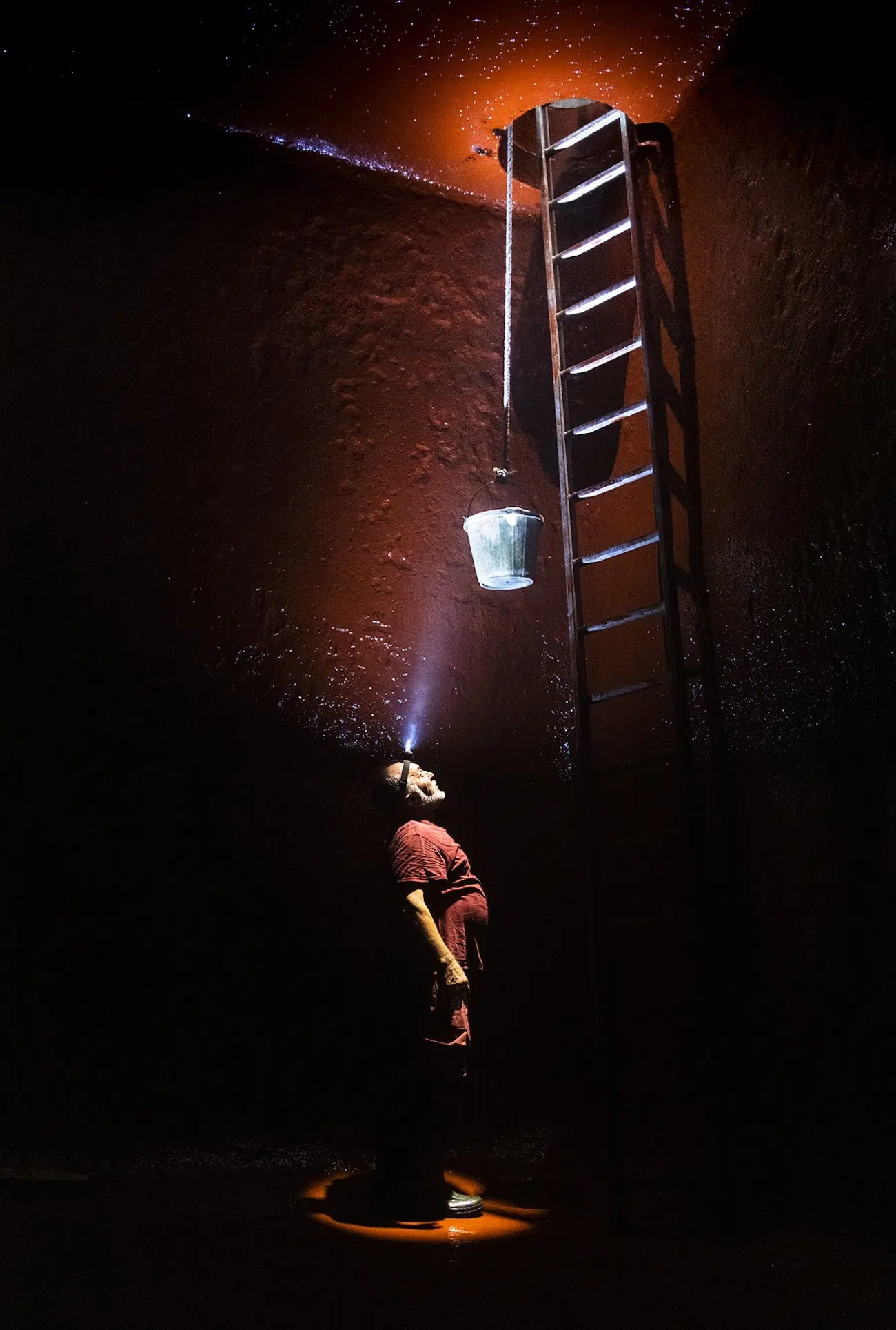 Person in a hole looking up a ladder with a light on their head