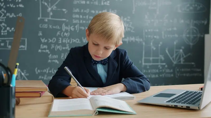 Elementary school student in a suit and bowtie writing in a notebook in a classroom
