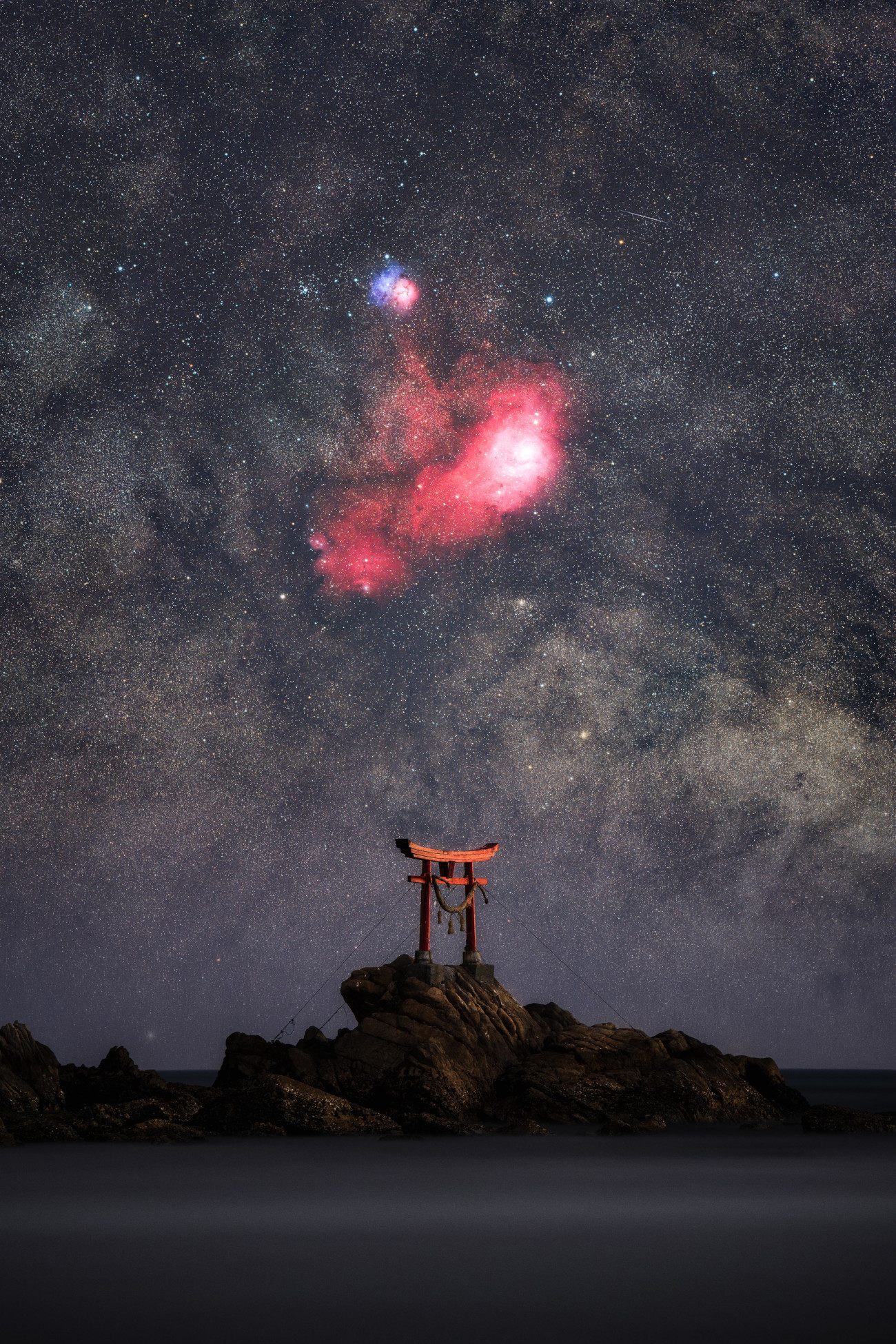 Skyscapes. "The Celestial Gate" © Yoshiki Abe (Japan). At Nata Beach, a solitary torii gate stands amid the waves. It marks a sacred site where one of the Japanese deities is said to have first descended to Earth. With the M8 and M20 nebulae shimmering in the background, the gate appears as a true portal to the heavens. Taken with a Sony ILCE-7RM5 camera, 200 mm, Foreground: f/5.6, ISO 320, 240-second exposure; Sky: f/4.0, ISO 3200, 34 x 60-second exposures; H-alpha: f/2.8, ISO 6400, 28 x 120-second exposures. Location: Kitsuki, Ōita Prefecture, Japan, 22 February 2026