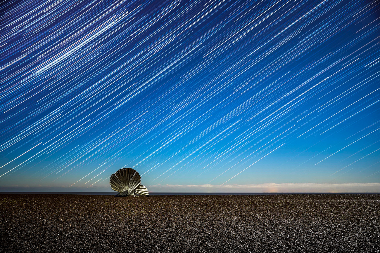 Skyscapes. "I Hear the Stars" © João Yordanov Serralheiro (Portugal). This image of star trails above the Scallop sculpture on Aldeburgh Beach, Suffolk, had been a long time in the making, as João Yordanov Serralheiro waited for the right night and conditions. It represents the quiet and peaceful feeling of sitting by the sea, listening to the waves come and go as time passes by –demonstrated here by the movement of the stars. Taken with a Sony Alpha 7 III camera, 35 mm f/2.8, ISO 400, 120 x 30-second exposures. Location: Aldeburgh Beach, Suffolk, England, 30 November 2025