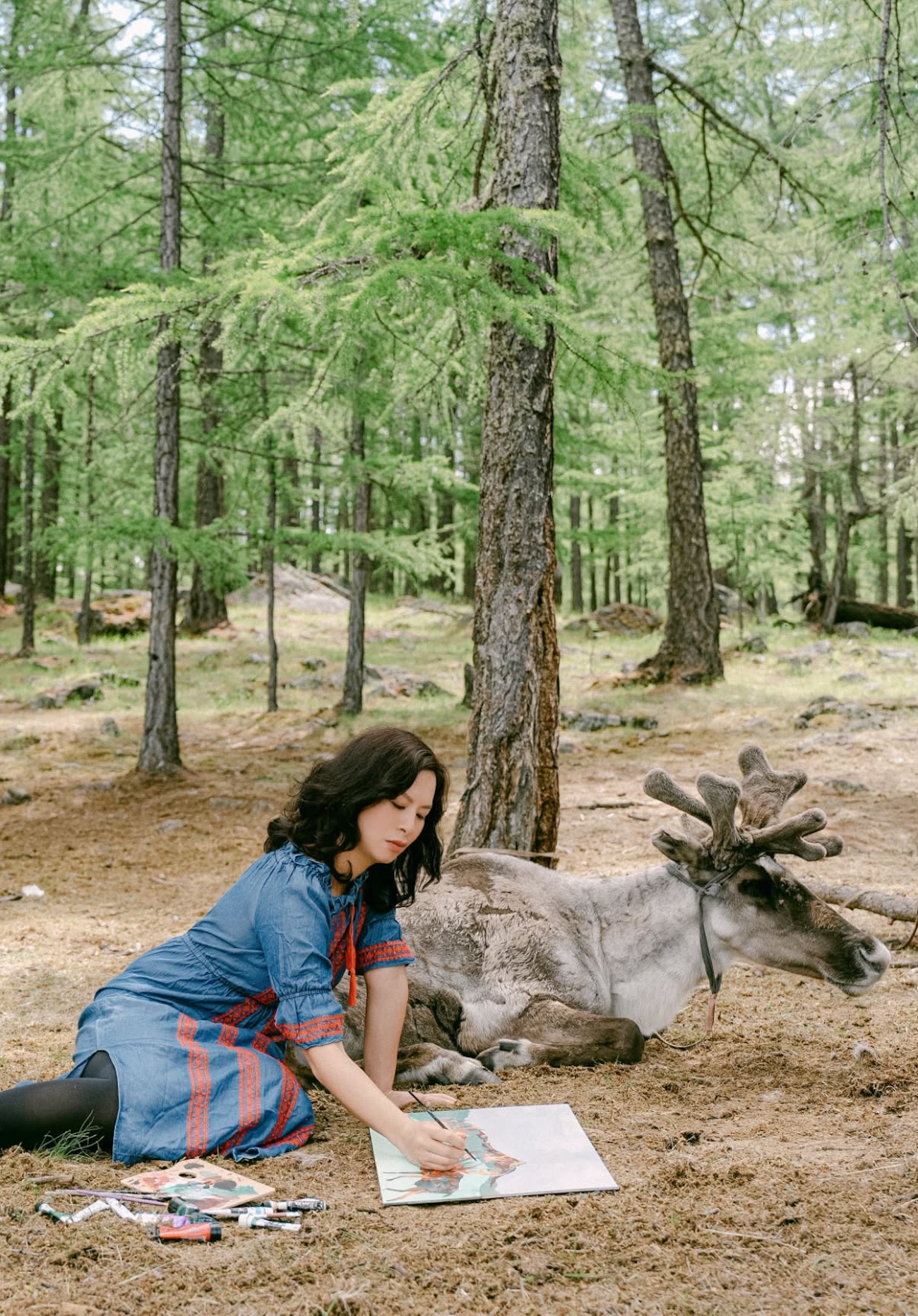 Photo of a woman painting next to an elk in the forest.